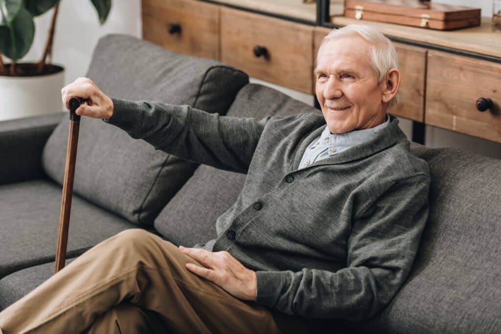 Senior man sitting on couch at home, smiling and holding cane